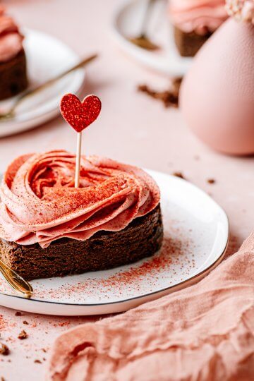 Erdbeer-Tonka-Brownie in Herzform auf einem Teller mit dekorativem Herzropper und rosafarbenem Toppinf auf einem romantisch gedeckten Tisch.